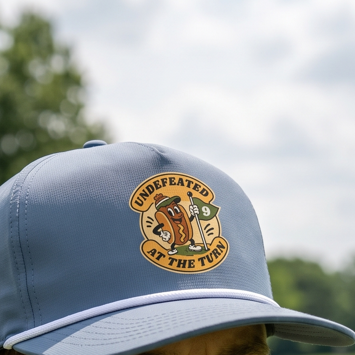 Close-up of the Undefeated at the Turn rope hat in Blue Jean, worn outdoors. The hot-dog-and-flag badge is printed flat into the twill weave, above a white braided rope.