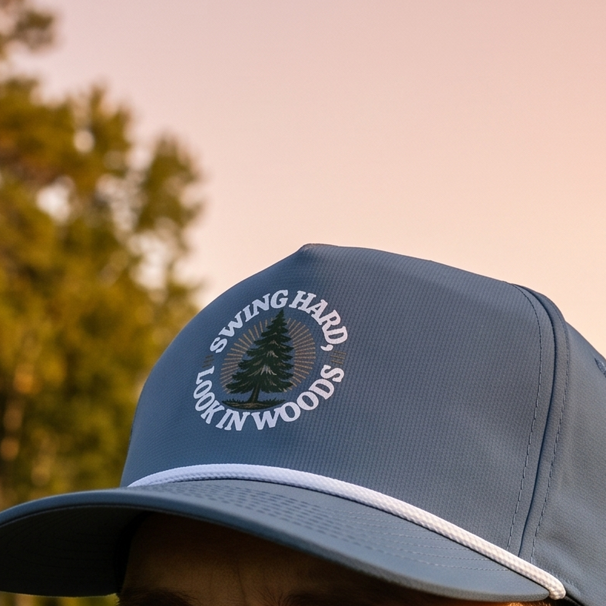 Close-up of the Swing Hard, Look in Woods rope hat in Blue Jean, worn outdoors at sunset. The circular pine-tree design is printed flat into the twill weave, above a white braided rope.