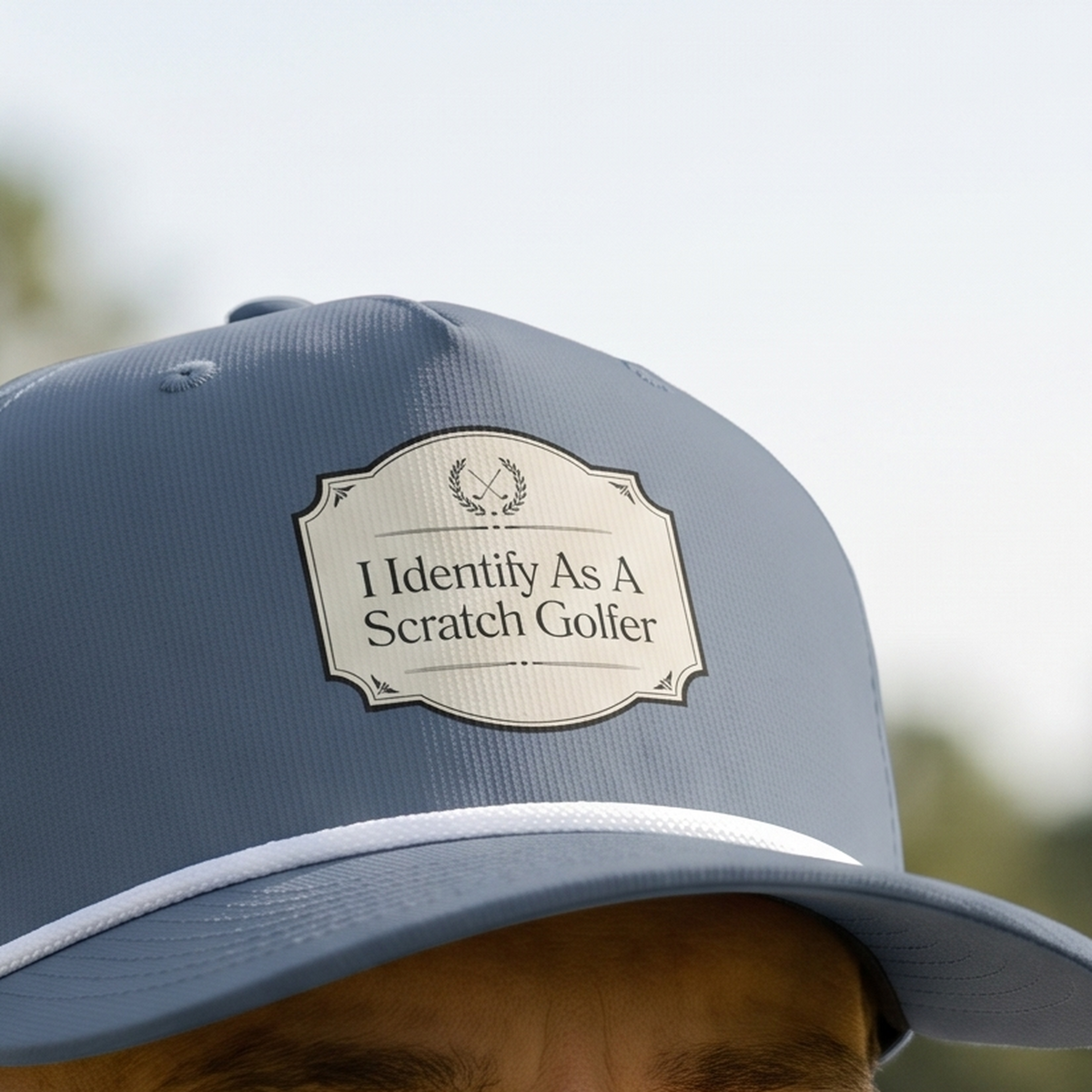 Close-up of the I Identify as a Scratch Golfer rope hat in Blue Jean, worn outdoors. The cream crest and serif lettering are printed flat into the twill weave, above a white braided rope.