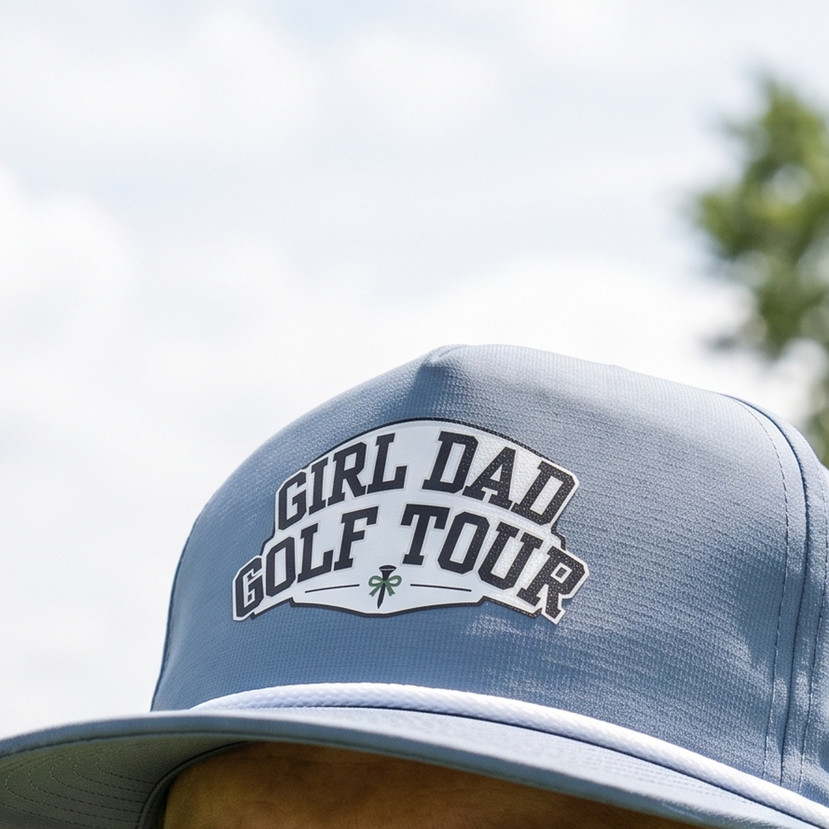 Close-up of the Girl Dad Golf Tour rope hat in Blue Jean, worn outdoors. The arched banner design is printed flat into the twill weave, above a white braided rope.