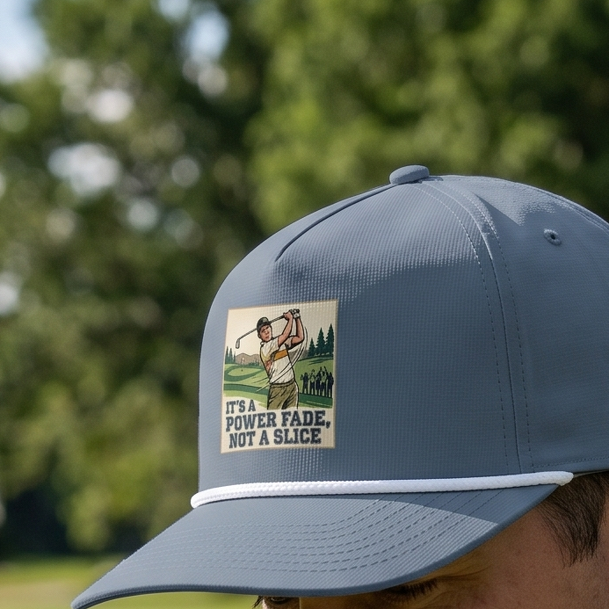 Close-up of the It's a Power Fade (Not a Slice) rope hat in Blue Jean, worn outdoors. The illustrated golfer panel is printed flat into the twill weave, above a white braided rope.