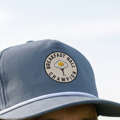 Close-up of the Breakfast Ball Champion rope hat in Blue Jean, worn outdoors. The circular fried-egg-on-a-tee design is printed flat into the twill weave, above a white braided rope.