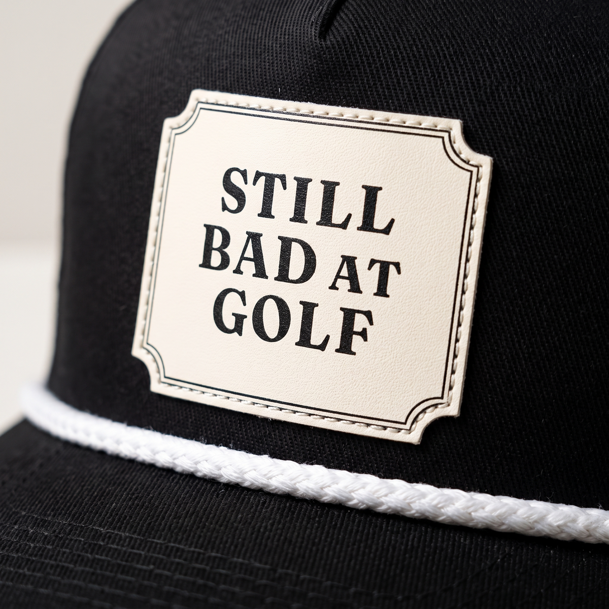 Close-up of the Still Bad at Golf rope hat in Black, showing the cream patch with its double-line border and the white braided rope across the bill.